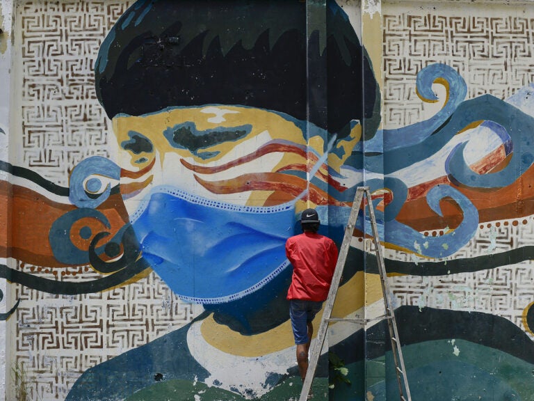 A street artist spray paints a protective face mask over an old mural featuring a Venezuelan Indigenous man