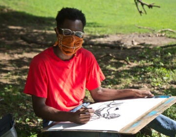 Nick Moncy, a young Black man, draws outside in a West Philadelphia park while wearing a mask