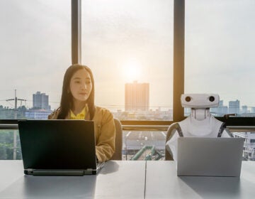 A young woman doing work on a computer, sitting next to a robot working on a computer
