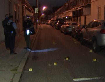 Philadelphia Police Department investigators stand on a sidewalk as bullet casings and evidence markers lay on the ground after a shooting