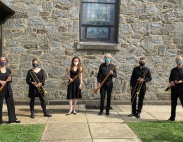 Musicians standing outside a stone building holding their instruments and wearing masks