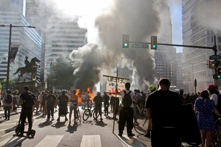 Smoke from burning police cars rises over protesters at Broad Street and JFK Boulevard. (Emma Lee/WHYY)