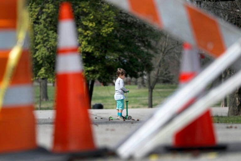 In this March 31, 2020 file photo, a child rides a scooter past barricades at an entrance to Tower Grove Park in St. Louis. (AP Photo/Jeff Roberson)