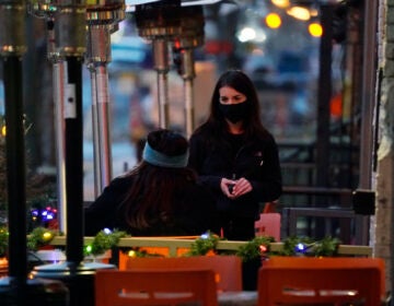 A waitperson wears a face mask while tending to a patron sitting in the outdoor patio of a sushi restaurant late Monday, Dec. 28, 2020, in downtown Denver. (AP Photo/David Zalubowski)
