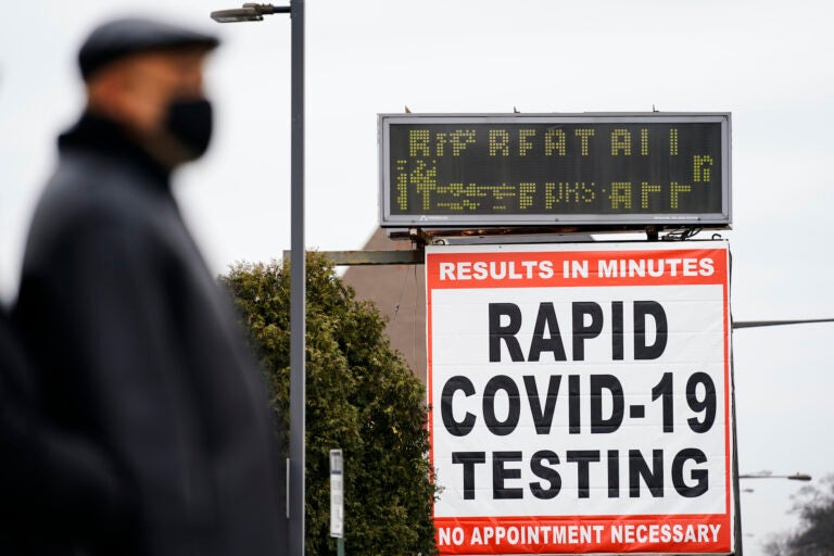 A person wearing a face mask as a precaution against the coronavirus stands near a sign advertising a rapid COVID-19 testing site in Philadelphia, Monday, Jan. 25, 2021. (AP Photo/Matt Rourke)