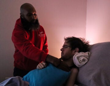 Ray Wall, a licensed practical nurse, positions Steven Sebastianelli in his bed. Wall visits Sebastianelli’s apartment every day to assist him in daily living activities. (Madison Karas/WHYY)