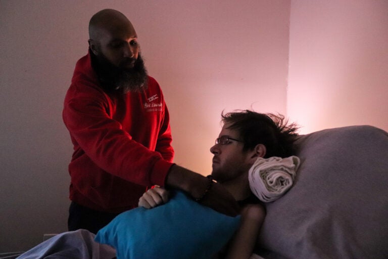 Ray Wall, a licensed practical nurse, positions Steven Sebastianelli in his bed. Wall visits Sebastianelli’s apartment every day to assist him in daily living activities. (Madison Karas/WHYY)