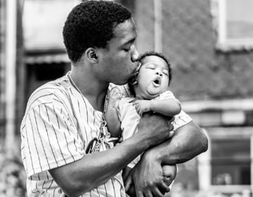 A photograph by Ken McFarlane showing a Black father kissing their baby in Movers & Makers: Made with Love