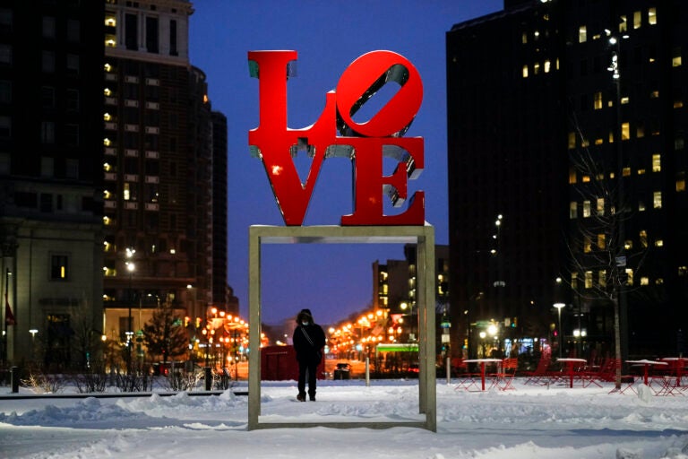 A person wearing a face mask as a precaution against the coronavirus walks during a winter storm near the Robert Indiana sculpture 