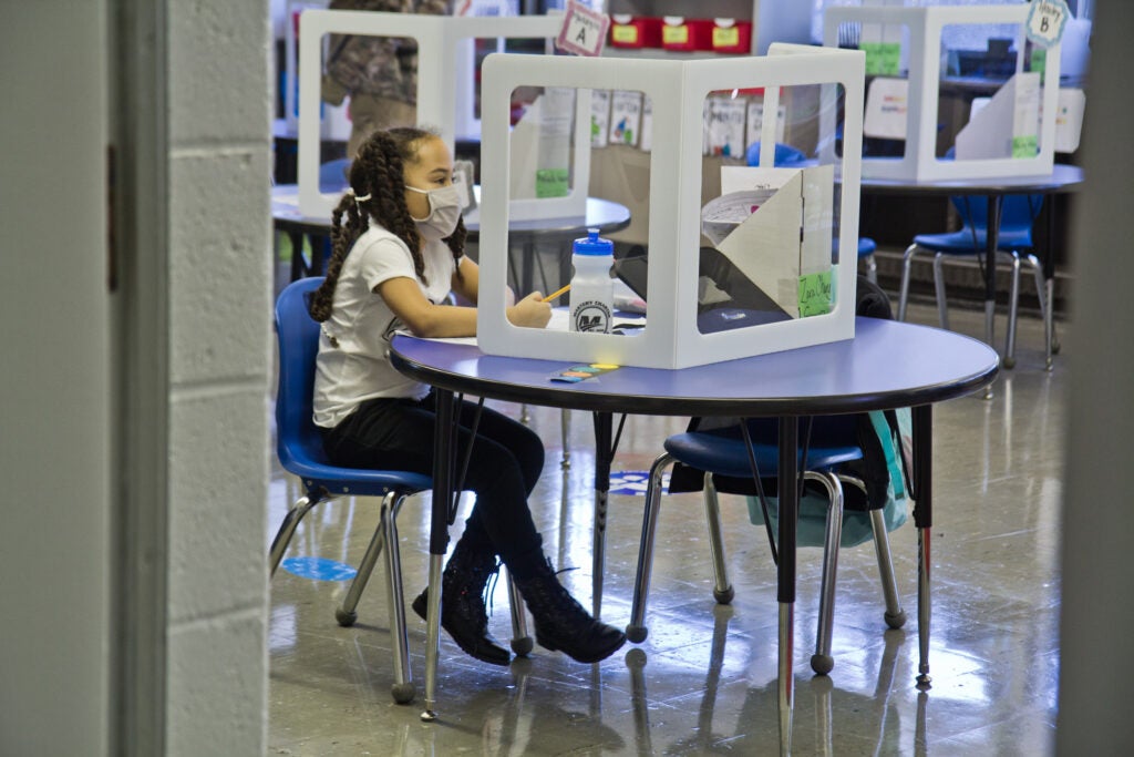 A student, wearing a face mask, sits at an isolated desk at school