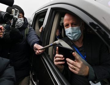 Peter Daszak holds up a phone from the passenger seat of a car while wearing a mask