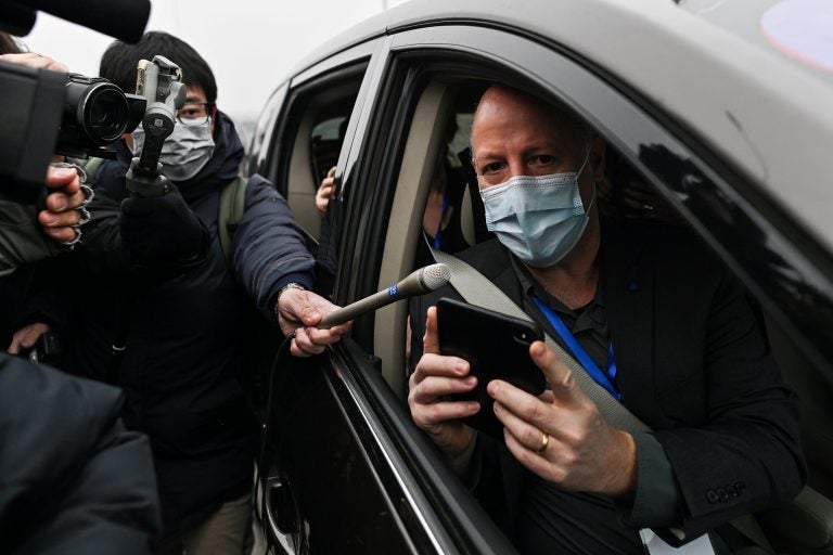 Peter Daszak holds up a phone from the passenger seat of a car while wearing a mask