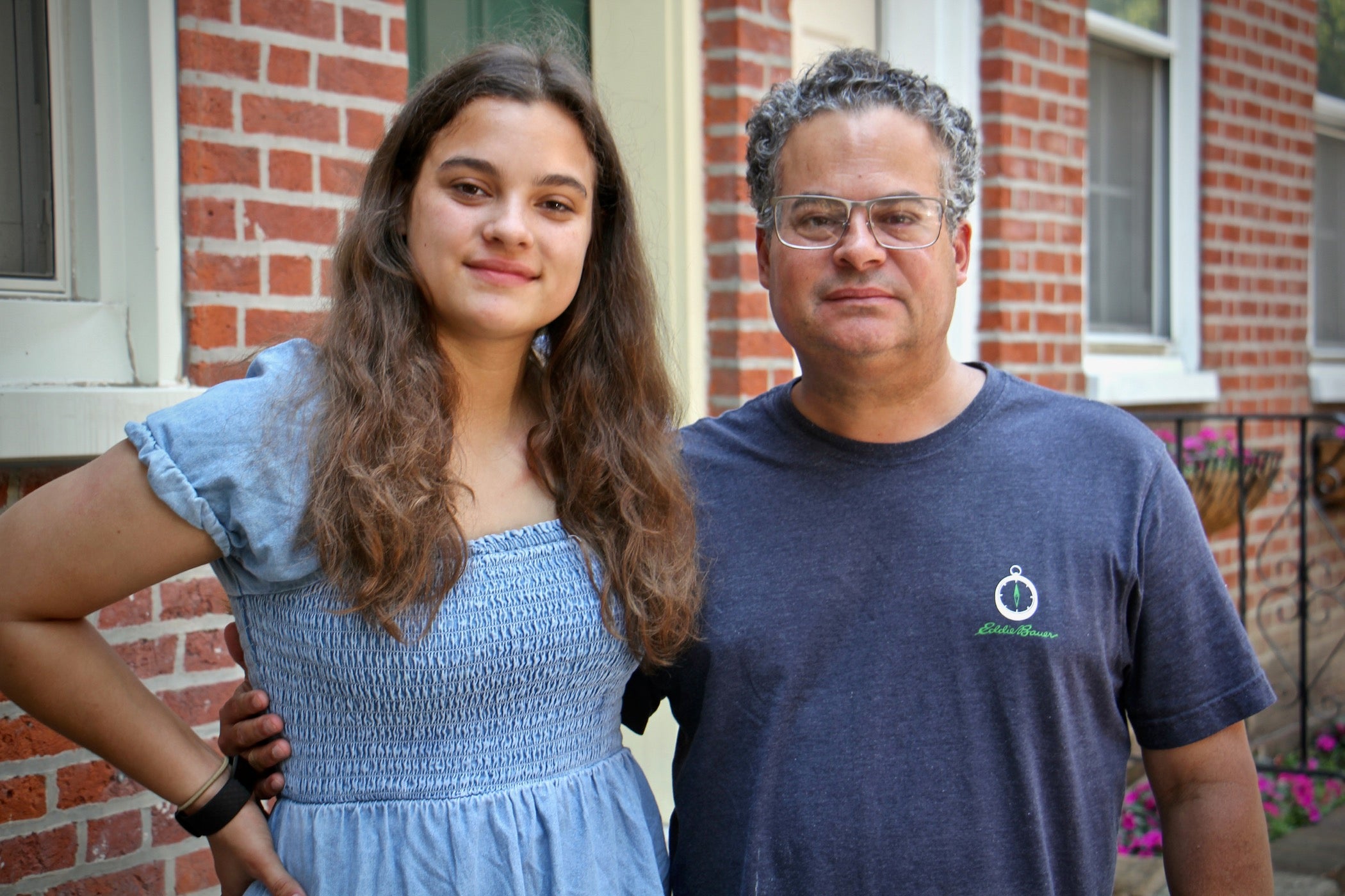A young mixed race woman stands next to her Latino father outside a brick home.