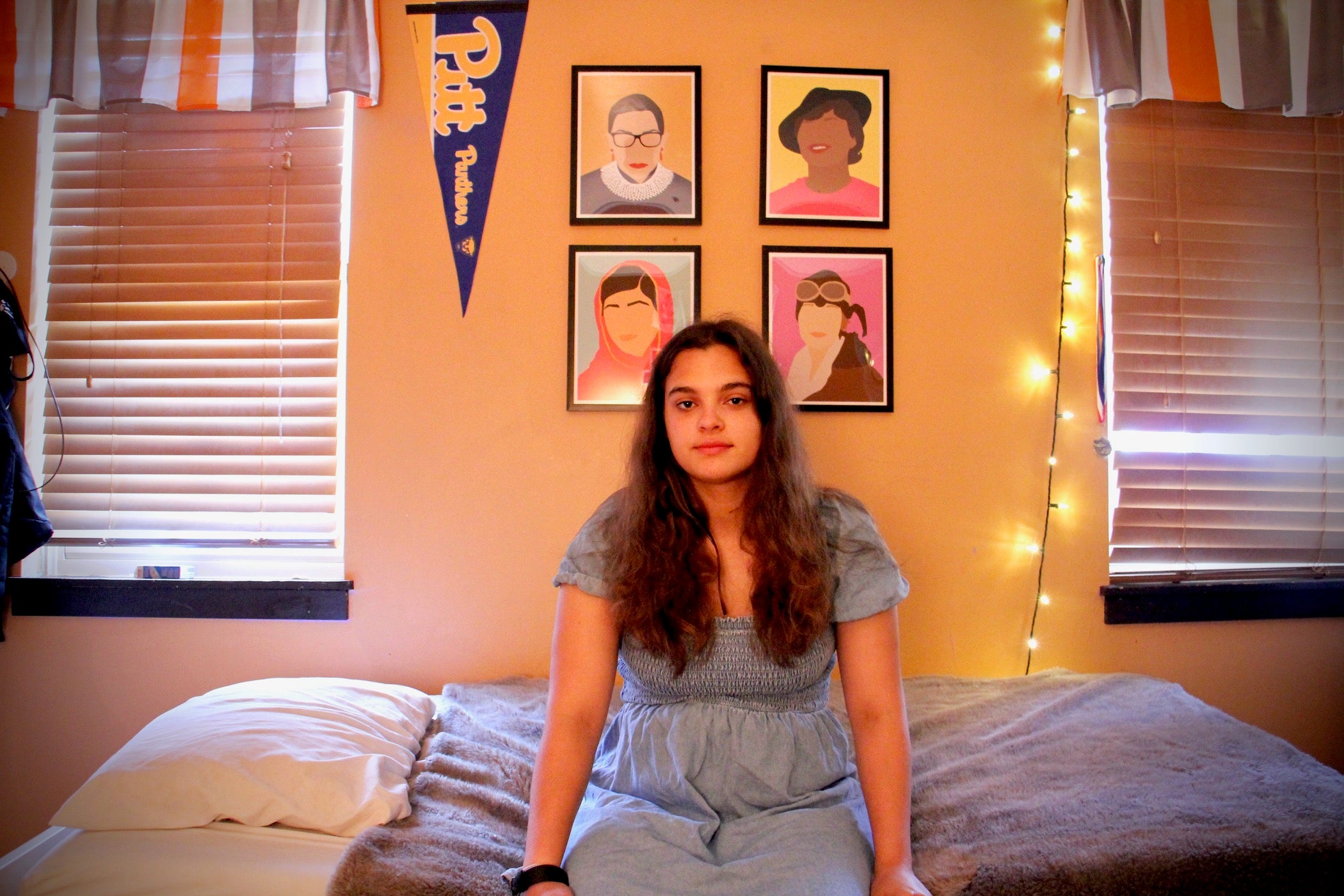 A mixed race teenager with long, brown hair sits on her bed. Four framed illustrations of Ruth Bader Ginsburg, Zora Neale Hurston, Malala Yousafzai and Amelia Earhart and a University of Pittsburgh pennant decorate the wall behind her.