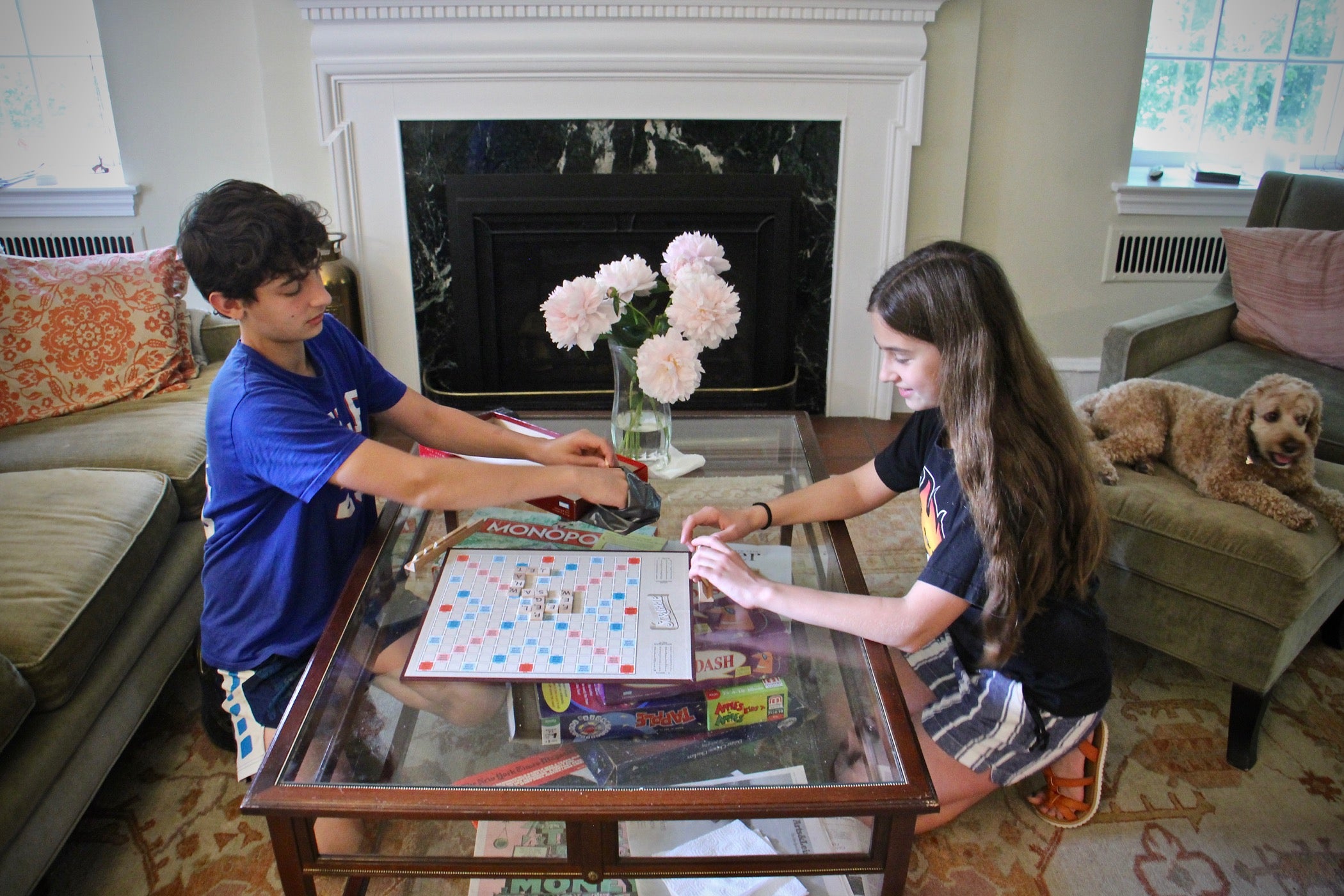 A teenage boy and his pre-teen sister kneel on the floor as they play Scrabble on a glass coffee table.
