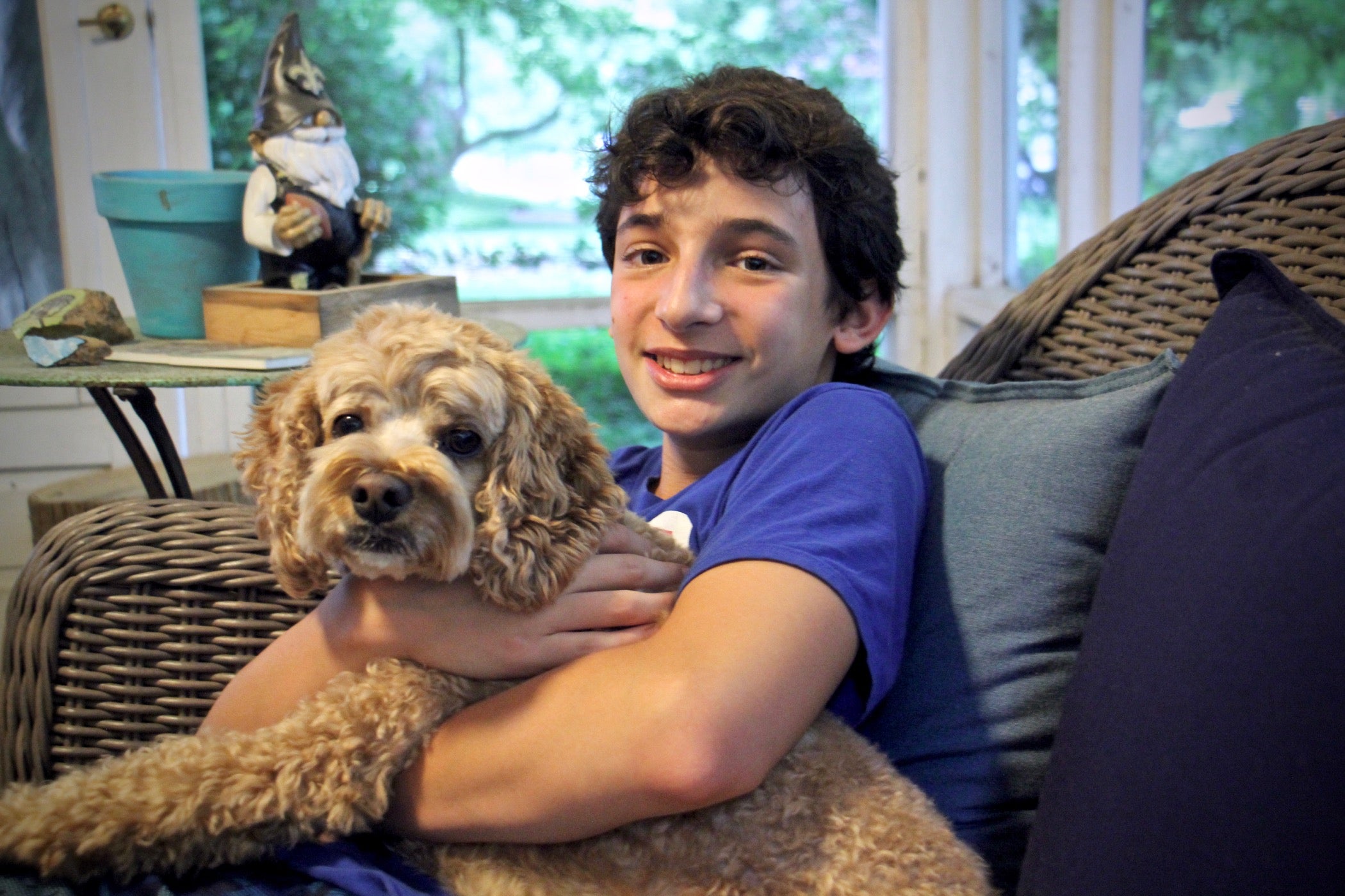 A white teenager with holds a medium-size dog with blond fur while sitting on a wicker chair.  