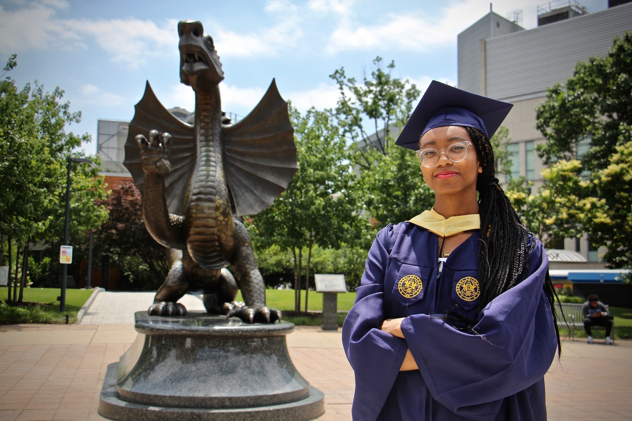 A young Black woman wearing a graduation cap and gown stands, arms crossed, next to a statue of Drexel University's dragon mascot.