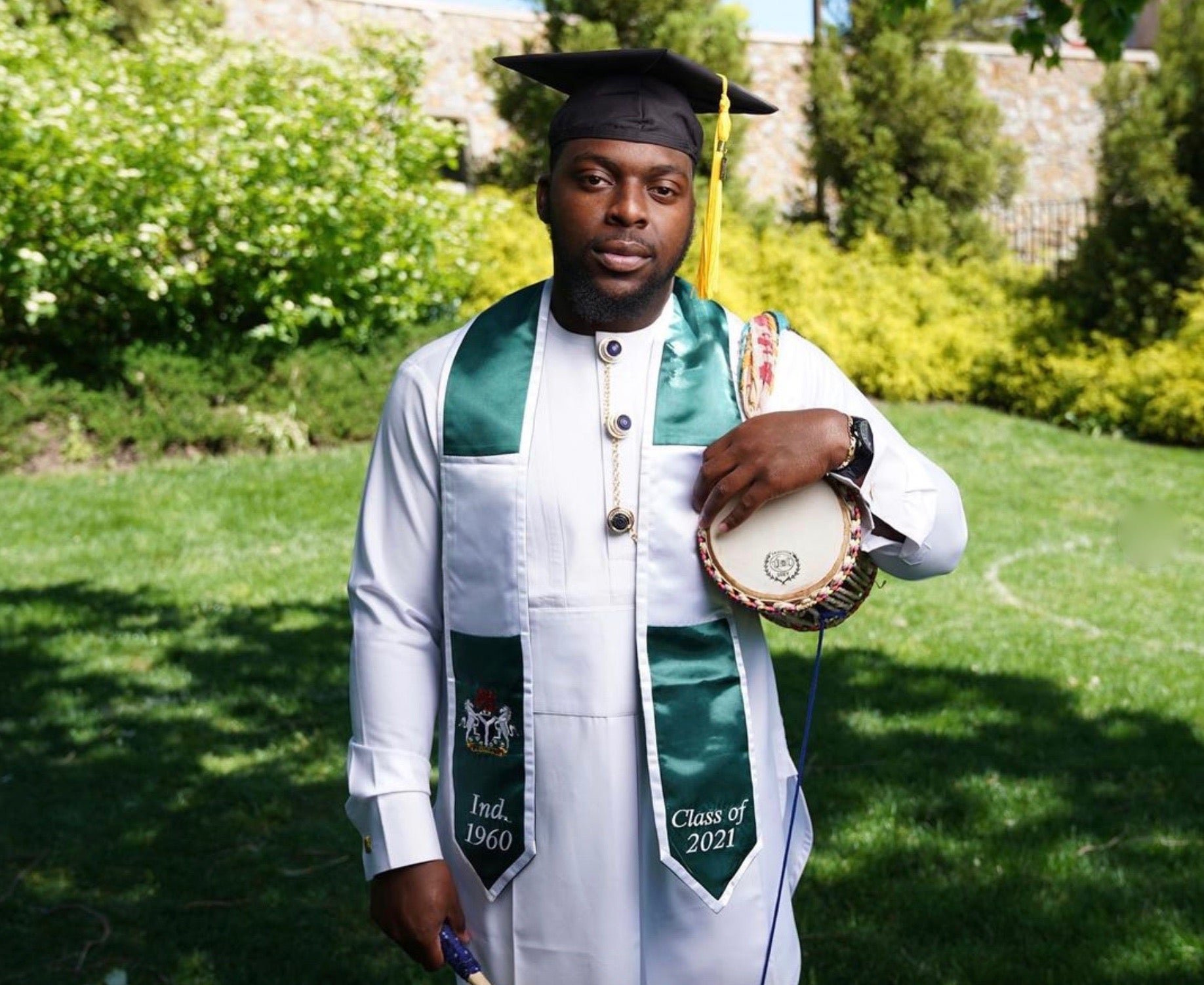 A young Black man, wearing a green and white graduation stole and black cap, holds a drum under his arm. Grass and bushes in the background.