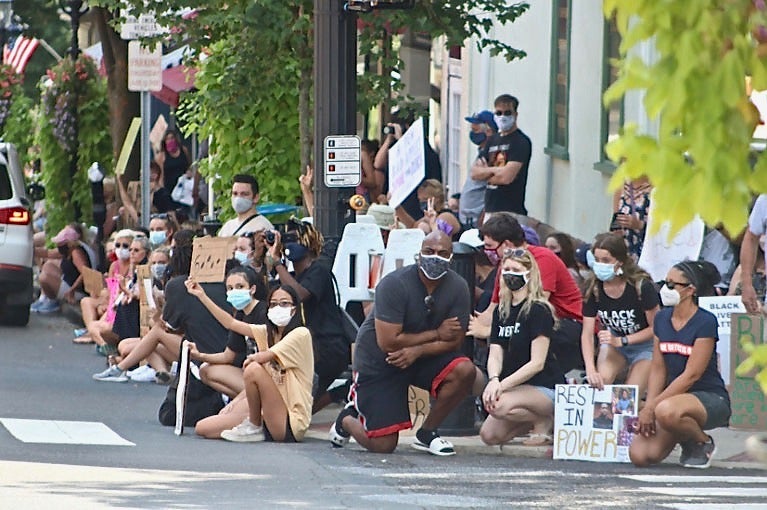 A crowd of people sit and kneel on the side of a street. The crowd is racially diverse. People are wearing masks and holding signs in support of Black Lives Matter.