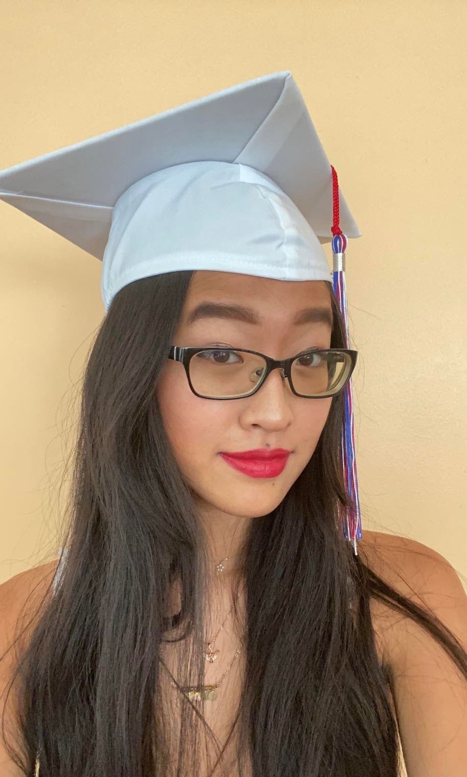 A young Chinese American woman with long brown hair and glasses wears a white graduation cap.