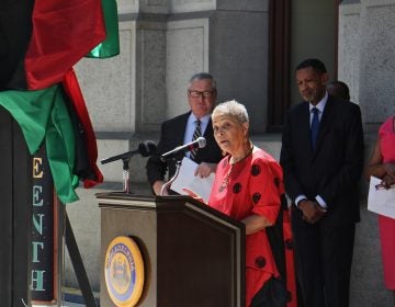 Helen Salahuddin speaks from a podium, with a Black Liberation Flag visible in the background