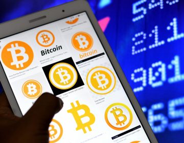 A hand holds up a phone with Bitcoin logos on it, against the backdrop of a screen with numbers on it