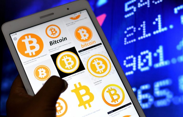 A hand holds up a phone with Bitcoin logos on it, against the backdrop of a screen with numbers on it