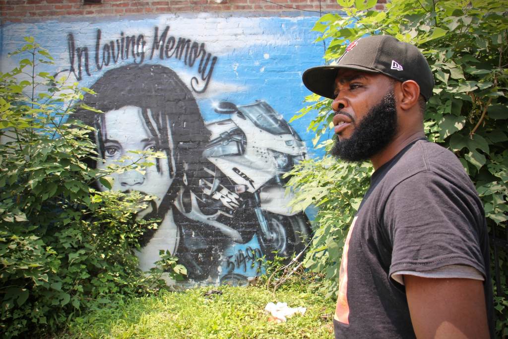 De'Wayne Drummond stands beside a mural dedicated to a motorcyclist who was killed at the intersection of 34th Street and Haverford Avenue