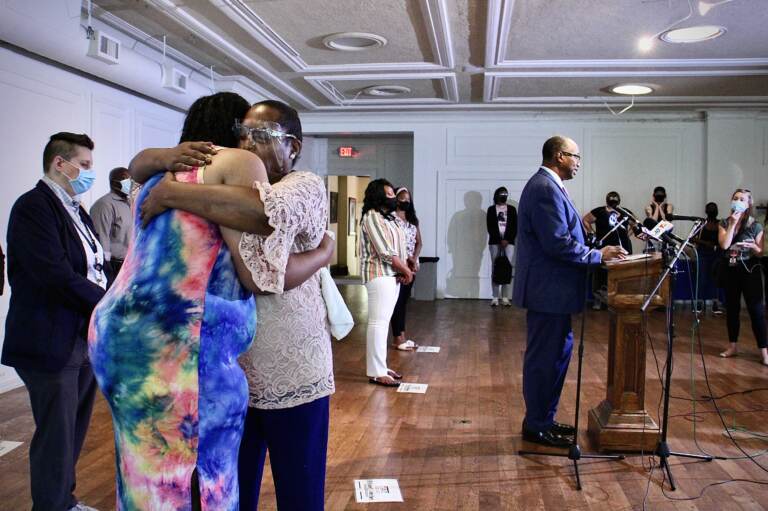 Rev. Andrea Harrington comforts Crystal Davis, with a speaker standing at a podium in the background