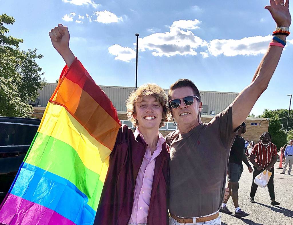 Bryce Dershem, wearing a pride flag, and his father hold their arms up in celebration