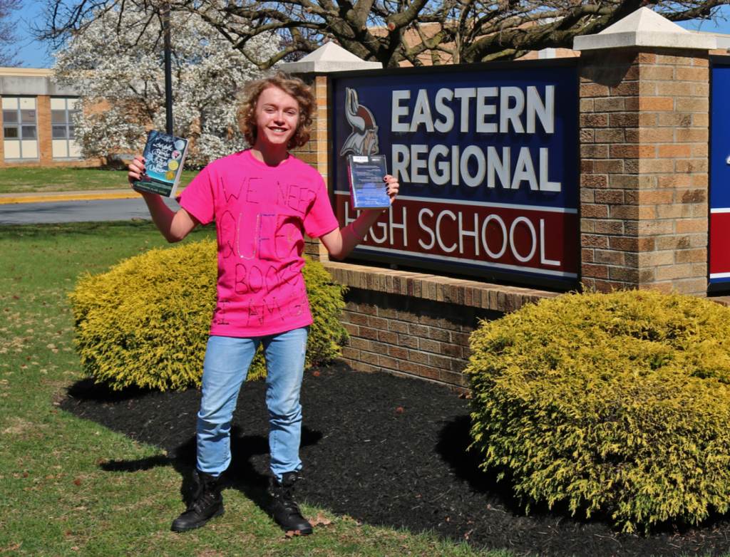 Bryce posses outside Eastern Regional High School wearing a T-shirt that says, "We need queer books, I am gay"