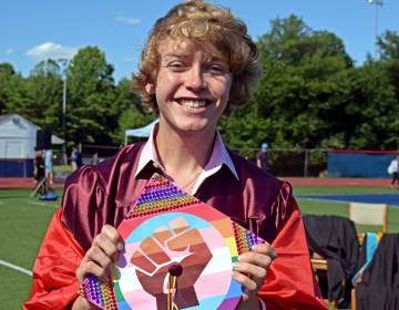Bryce Dershem holds up his graduation cap, which features a fist on top of a trans pride flag
