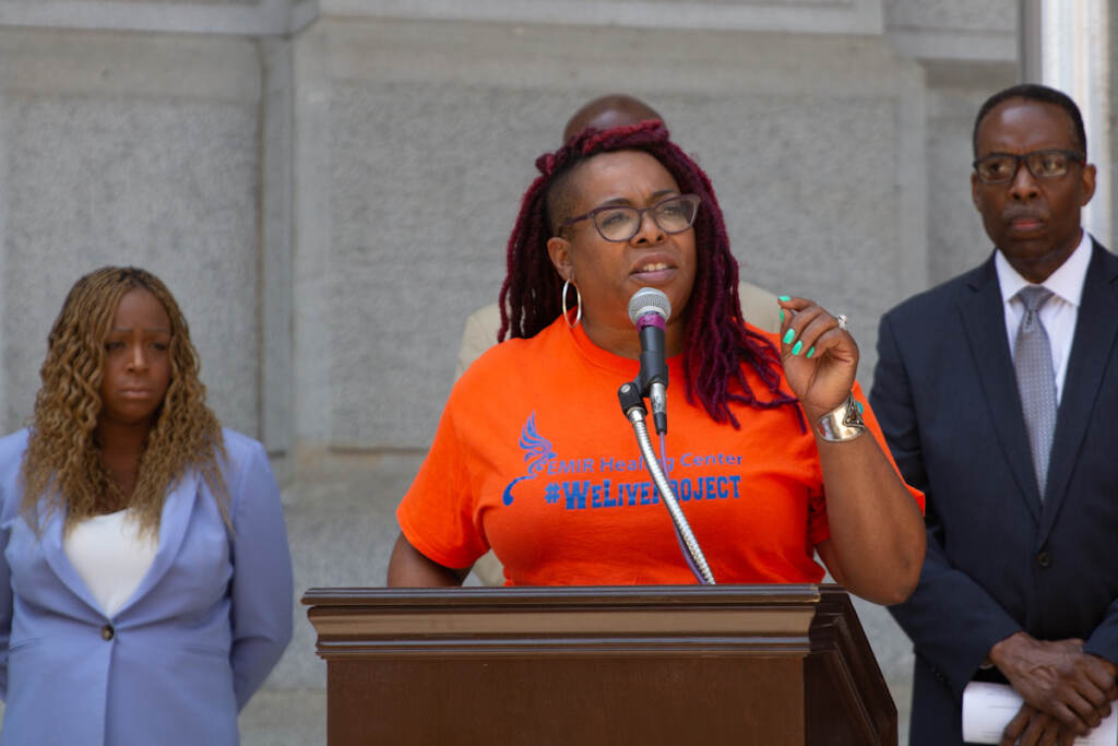 Chantay Love gestures while speaking from a podium, with elected officials behind her