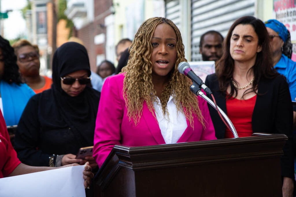 speaks from a podium at a press conference, with elected officials behind her