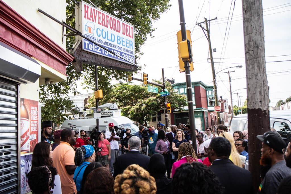 Philadelphia elected officials and community leaders gather outside a convenient store for a press conference, with a crowd in the foreground