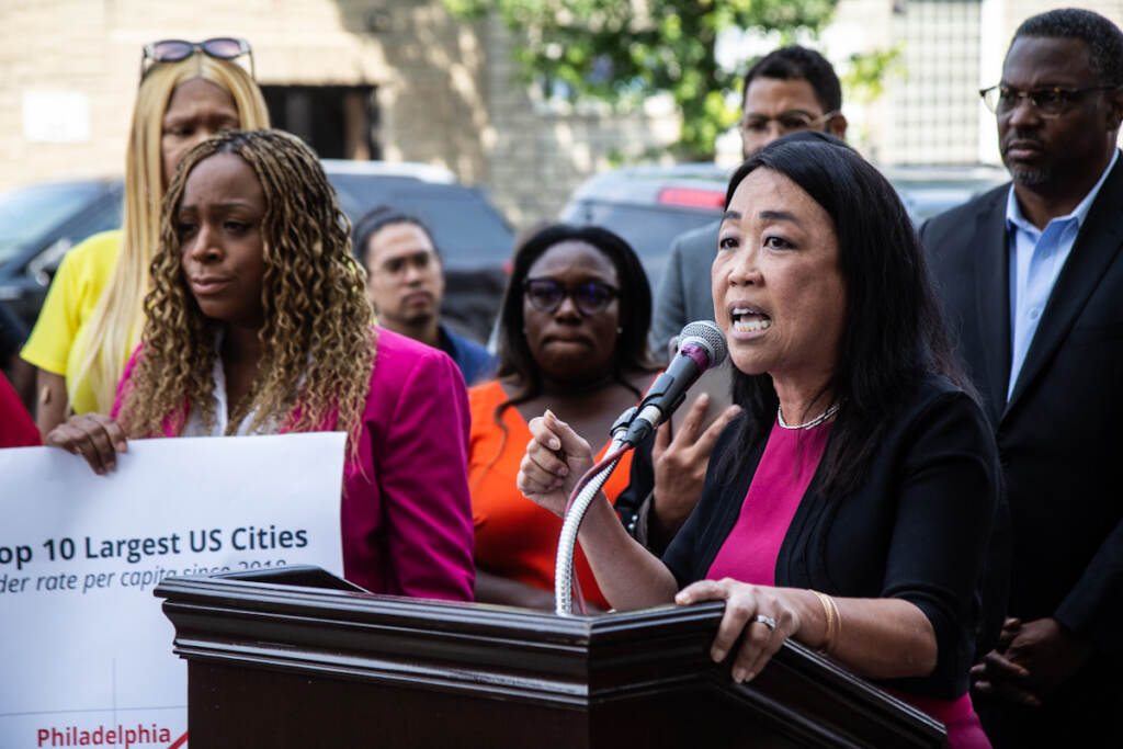 Helen Gym speaks from a podium at a press conference, with elected officials behind her