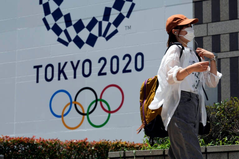 A woman wearing a protective mask walks with a baseball cap to shield from the sun walks in front of a Tokyo 2020 display