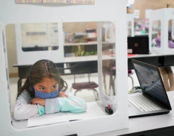 Londyn Vargas, seen behind a see-through partition, puts her head in her arms while sitting at a desk in her classroom