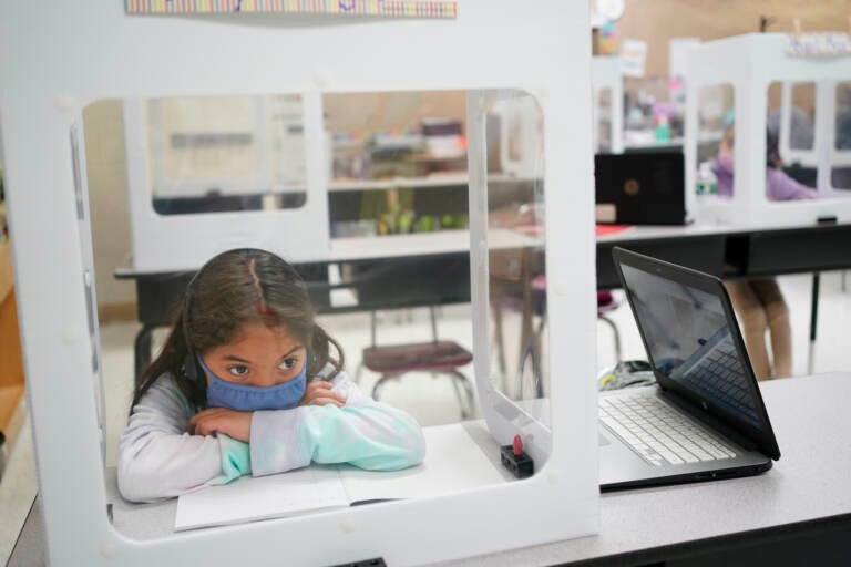 Londyn Vargas, seen behind a see-through partition, puts her head in her arms while sitting at a desk in her classroom