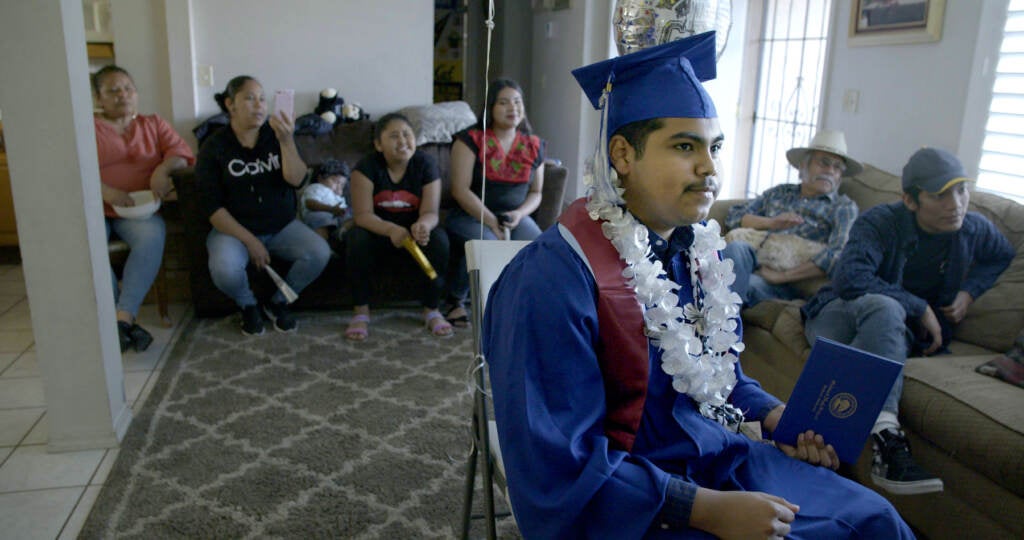 A still from the documentary "Homeroom" shows a person wearing a graduation gown, with people sitting on a couch behind them