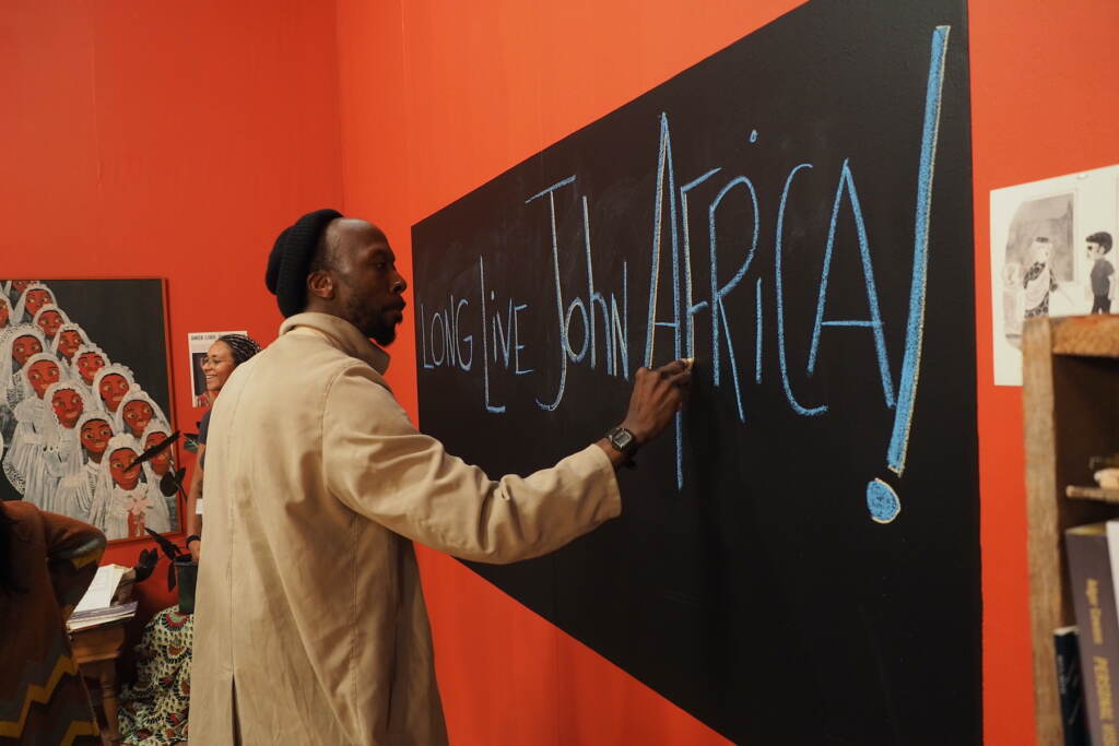 A still from "The Inheritance" shows a man at a chalkboard, with the words "Long live John Africa" written