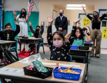 Kindergarteners raise their hands while wearing face masks in a classroom