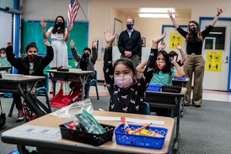 Kindergarteners raise their hands while wearing face masks in a classroom