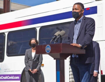 A man stands at a podium with a bus visible behind him.