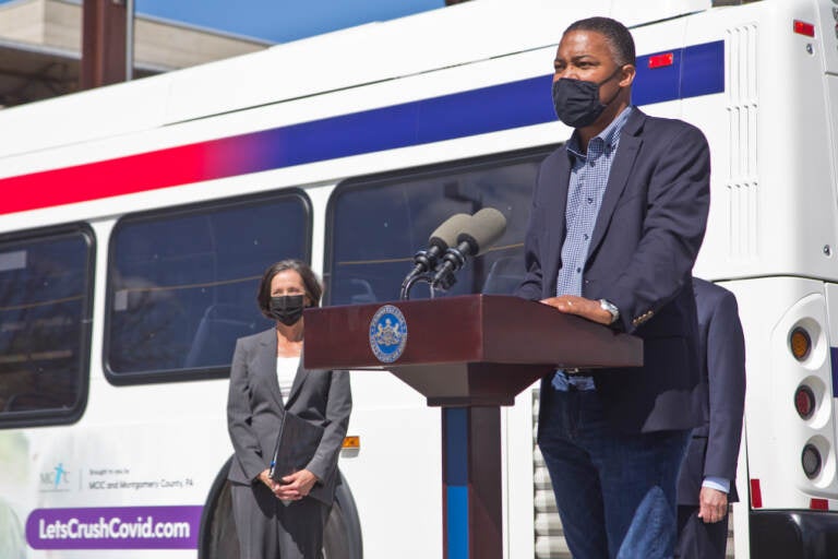 A man stands at a podium with a bus visible behind him.