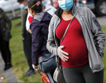 A pregnant woman wearing a face mask and gloves holds her belly as she waits in line for groceries