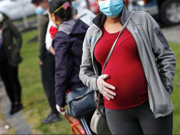 A pregnant woman wearing a face mask and gloves holds her belly as she waits in line for groceries