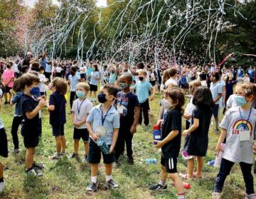 Kids, wearing masks, are surrounded by confetti outside during a ceremony