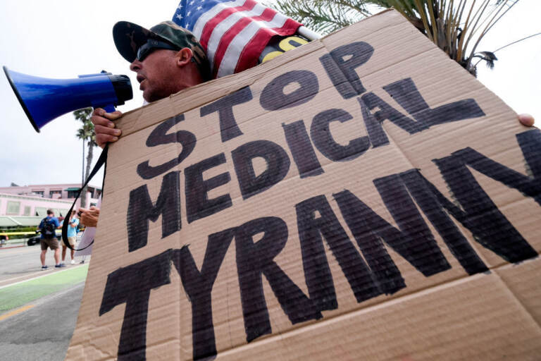 An anti-vaccination protester holds a sign and a flag as he takes part in a rally against Covid-19 vaccine mandates, in Santa Monica, California, on August 29, 2021. (Photo by RINGO CHIU / AFP) (Photo by RINGO CHIU/AFP via Getty Images)