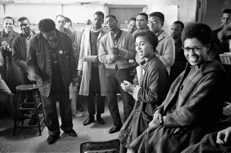 James Forman leads singing in the SNCC office on Raymond Street in Atlanta, (from left) Mike Sayer, MacArthur Cotton, Forman, Marion Barry, Lester McKinney, Mike Thelwell, Lawrence Guyot, Judy Richardson, John Lewis, Jean Wheeler, and Julian Bond, 1963, Danny Lyon